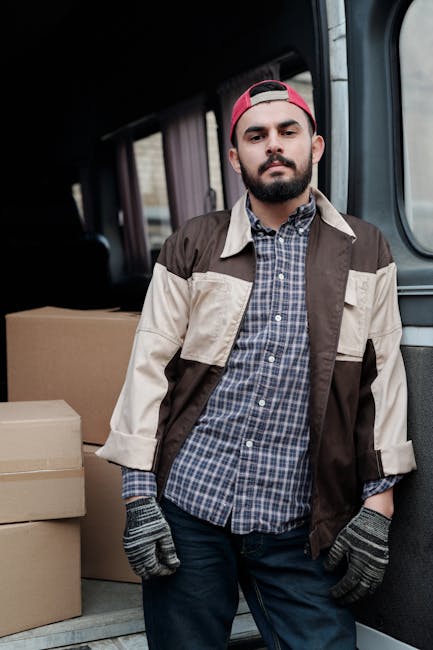 A man with a beard and wearing a red baseball cap, a brown and beige jacket over a blue checkered shirt, and grey work gloves, stands inside the back of a van during a house removal or furniture transport. The interior of the van features dark upholstery, and in the background, there are cardboard boxes of various sizes prepared for moving, some stacked on the floor. The van appears to be in a driveway or outdoor loading area, with natural light illuminating the scene. This image illustrates the packing and loading process involved in home relocation, with an emphasis on careful handling of belongings, consistent with services provided by Man with Van South Hackney, a company specialising in removals and local moves around Dalston and South Hackney.