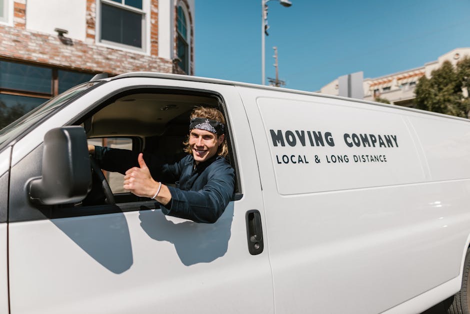A smiling young man with long hair, wearing a black headband and a navy blue shirt, is sitting in the driver’s seat of a white moving van with the company's name and services printed on the side, including 'MOVING COMPANY' and 'LOCAL & LONG DISTANCE.' The van is parked outside a residential building with brick and glass exterior walls under a clear blue sky. Inside the vehicle, the driver is visible through the open window, giving a thumbs-up gesture, while the interior shows the dashboard and steering wheel. The scene suggests a home relocation or furniture transport process, with the man likely preparing for a move or guiding the loading process of household items, such as cardboard boxes, furniture, or appliances, onto the vehicle. The setting emphasizes professional local and long-distance removal services by Man with Van South Hackney as part of a house moving or packing and moving operation.