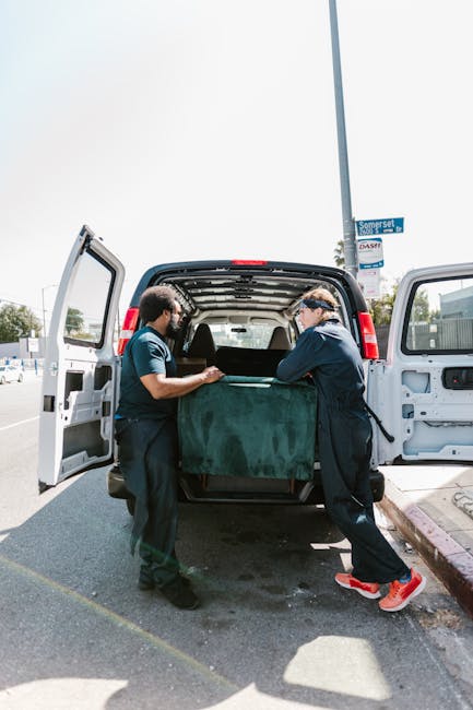 A man with a beard and wearing a red baseball cap, a brown and beige jacket over a blue checkered shirt, and grey work gloves, stands inside the back of a van during a house removal or furniture transport. The interior of the van features dark upholstery, and in the background, there are cardboard boxes of various sizes prepared for moving, some stacked on the floor. The van appears to be in a driveway or outdoor loading area, with natural light illuminating the scene. This image illustrates the packing and loading process involved in home relocation, with an emphasis on careful handling of belongings, consistent with services provided by Man with Van South Hackney, a company specialising in removals and local moves around Dalston and South Hackney.