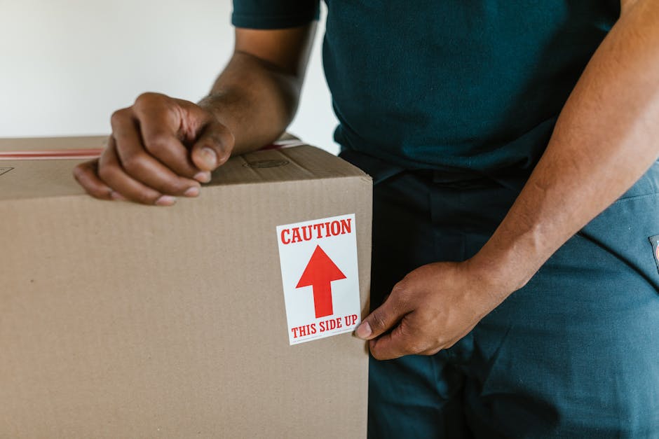 Close-up of a person wearing a dark teal shirt and dark trousers, handling a large cardboard box during a home relocation process. The individual is attaching a red and white caution sticker that reads 'CAUTION THIS SIDE UP' with an upward arrow, indicating proper orientation for moving. The box is positioned near a doorway or entryway with visible beige carpet flooring, and appears to be part of the packing and moving preparations. In the background, a glimpse of the interior environment suggests an indoor setting, with the focus on safe handling and transportation of packed items. This image illustrates the careful packing and loading procedures typical of furniture transport and house removals services provided by Man with Van South Hackney, supporting professional logistics for residential relocations.