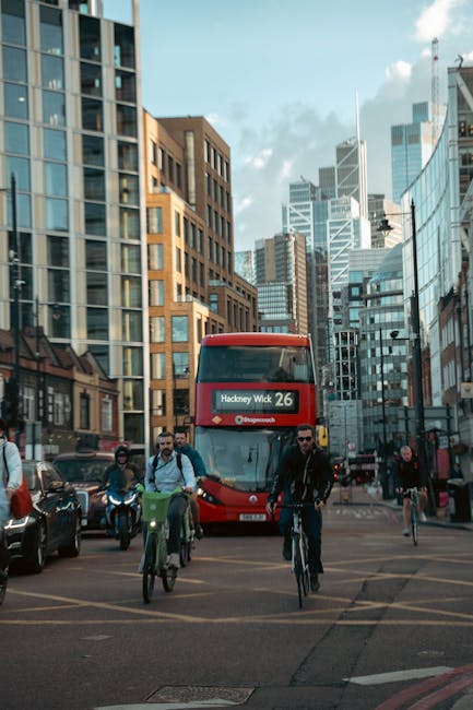 In the foreground, a group of cyclists, including a person in a black jacket and sunglasses, are riding bicycles across a busy city street during daylight hours. Behind them, a red double-decker bus displaying 'Hackney Wick 26' as its route is approaching, with various pedestrians walking along the sidewalk and waiting at the crosswalk. The street is lined with modern high-rise buildings made of glass and concrete, reflecting the bright sky with scattered clouds. To the left of the bus, cars are parked along the curb, and a woman with a red bag is visible walking on the sidewalk. The scene captures a typical urban environment with active transportation and urban architecture, illustrating a City of London setting conducive to house removals and transport services provided by Man with Van South Hackney.