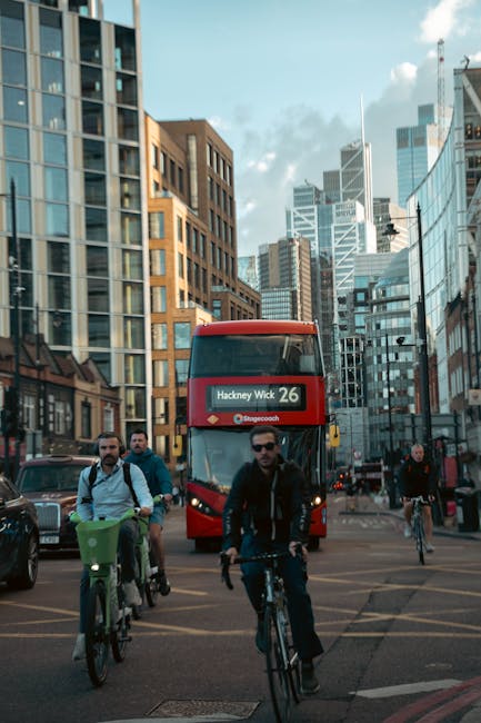 Two black London taxis with illuminated round headlights are parked side by side on a city street, facing the camera. Behind them, several cars are lined up along the curb, with a red double-decker bu