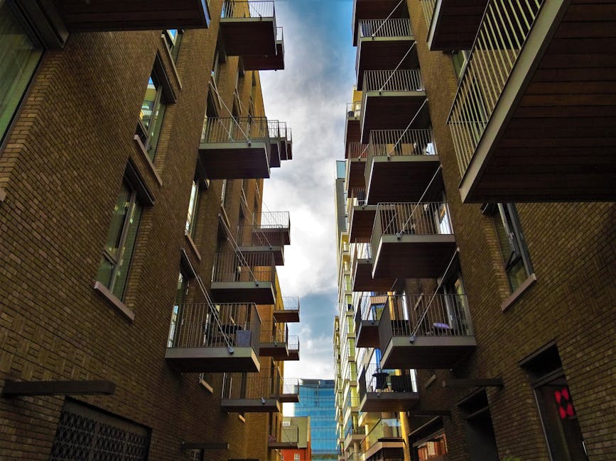 View looking up between two adjacent brick apartment buildings with multiple small balconies featuring metal railings, some with visible furniture or items, and large windows on each floor. The balconies are positioned at different levels, creating a vertical corridor effect. In the background, a portion of a modern glass building and a cloudy sky are visible. The scene is set during daylight, with natural light illuminating the buildings' facades. This image captures a typical urban residential environment suitable for house removals and furniture transport, emphasizing the narrow access and vertical living spaces often encountered in city relocations, as managed by Man with Van South Hackney.