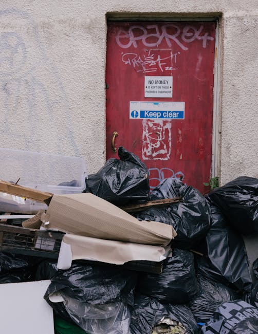 A red metal door with graffiti and multiple signs including 'No Money' and 'Keep Clear' is situated in an exterior wall of a building with a rough, textured surface. In front of the door, there is a p
