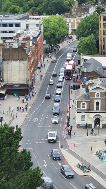 An aerial view of a busy street in Homerton E8 during daylight, with a line of vehicles including a red double-decker bus, cars, and vans moving along the road. On the left side, pedestrians are walking on the sidewalk next to a row of shops and buildings, while on the right, there are parked bicycles and a small structure with a pitched roof. Large, leafy green trees line both sides of the street, providing partial shade. The scene captures the typical urban environment associated with home relocation activities, with visible traffic flow and foot traffic as part of the moving logistics process supported by Man with Van South Hackney's removal services.