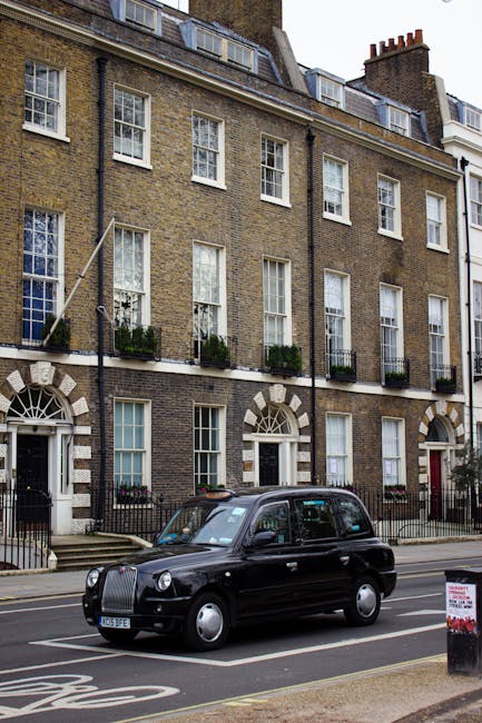 A black London taxi cab parked on the street in front of a multi-storey brick residential building with large sash windows and small black iron balconies adorned with potted plants. The building has a traditional Georgian architectural style with white stone accents around the arched doorway and window frames. The taxi is positioned near the curb on a paved road, with a roadside sign visible in the foreground. The scene suggests a typical urban setting suitable for house removals in South Hackney, with the vehicle possibly used for furniture transport or home relocation services provided by Man with Van South Hackney, supporting the home packing and moving process.