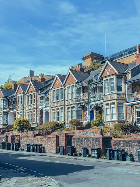A row of Victorian-style terraced houses with ornate bay windows, decorative iron railings, and brick facades, situated on a residential street in South Hackney. The houses are elevated above the pavement with landscaped front gardens bordered by low brick walls. Several black wheelie bins are lined up along the curb, and the street features a clear, blue sky overhead, indicating daytime. In the foreground, part of a parked van with open doors and wrapping materials can be seen, suggesting the process of home relocation or furniture transport. The scene reflects a typical London neighbourhood involved in packing and moving activities, with visible elements of house removals and logistical support by [COMPANY_NAME] for a residence transfer to Mare Street, as referenced in the page title.
