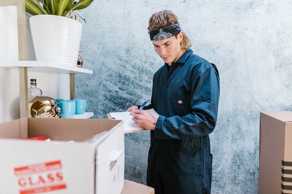 A young male mover wearing a black work jumpsuit and a black and white bandana is inside a room with a textured light blue wall, preparing for a home relocation. He is holding a clipboard and a pen, reviewing documentation related to the packing and moving process. In front of him are cardboard boxes, one of which is open and contains red packing paper, and other boxes sit on a shelf behind him. On the shelf, there are decorative items including a large white ceramic pot with a plant, a glass bottle, and teal-colored mugs. The environment is well-lit, highlighting the packing and preparation stage of furniture transport. This scene depicts the careful planning involved in house removals and the logistical coordination by [COMPANY_NAME] as part of a professional packing and moving service targeting clients in the Victoria Park and Hackney areas.