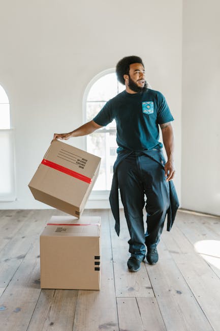 A man with dark curly hair and a beard is inside a well-lit room with white walls and large arched windows, engaged in a home relocation task. He is wearing a navy blue T-shirt with a small logo on the chest, dark trousers, and has a jacket tied around his waist. He holds a cardboard box, secured with red tape, in his right hand, preparing to place or move it onto another box positioned on the wooden floor. The room features wooden flooring, and the sunlight streaming through the windows illuminates the space, highlighting the moving process. This scene reflects furniture transport and packing activities typical of professional removals offered by [COMPANY_NAME], supporting services such as house moves and furniture transport during a residential relocation as outlined in the page titled 'Victoria Park fragile item removals expert tips for Hackney, SOUTH HACKNEY'.
